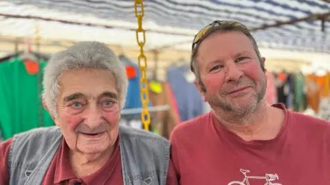 George Carden/BBC Andrew Sleeman and Alf Franks standing in a clothes stall with clothes hanging behind them. Both are wearing maroon tops and Alf has a grey denim gilet and grey hair. 