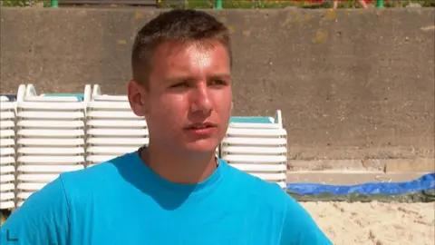 Vittorio Macari is pictured in a light blue T-shirt. He is standing in front of stacks of beach loungers on the beach and there is a brown granite wall in the background