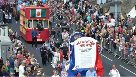 BBC A view of the 70th Liberation Day cavalcade when floats were last included. One float is designed to look like an old red Guernsey tram and another is red white and blue and says Occupation Nights on it 