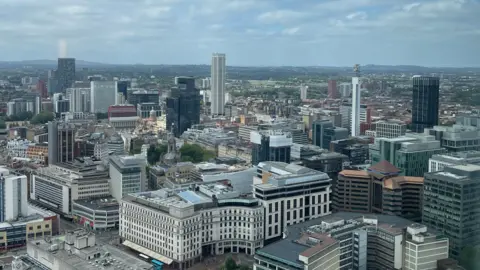 An aerial view of Birmingham city centre taken from high up in One Eastside. Rooftops, offices and residential buildings stretch out into the distance under a cloudy blue sky. The BT Tower and other well-known skyscrapers are visible on the horizon.