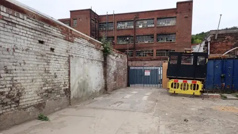 Defra A narrow, partly enclosed yard is bordered by weathered brick walls and industrial buildings. In the foreground, a cracked concrete and gravel surface leads to a locked metal security gate at the far end of the yard. 