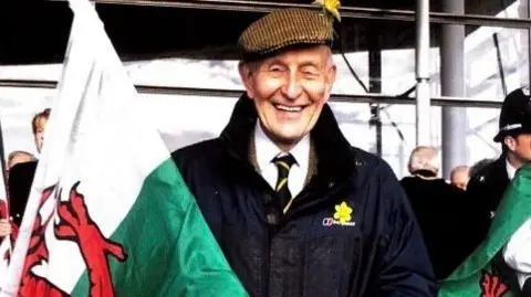 Family Photo Dewi Bowen wearing a white shirt and tie and navy coat looking at the camera with a large Welsh flag in his hand. 