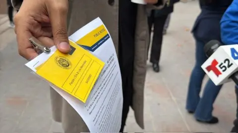 Christian Garcia is seen holding up a bright yellow badge clipped to several sheets of paper about the Menendez brothers case. The badge reads "People v Erik & Lyle Menendez" 