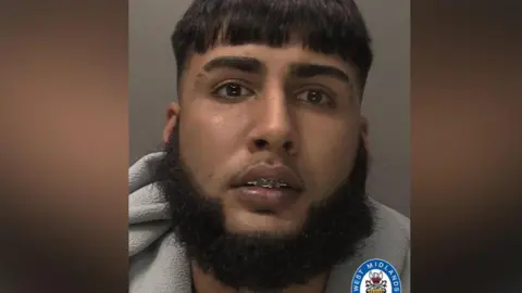 A police mug shot of Gurpreet Randhawa. He has dark hair and a beard and what looks like braces on his teeth. 