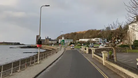 Google A road with water on the left and houses on the right. Lampposts line the water side of the road. There is a pavement on that side as well and trees can be seen in the distance.