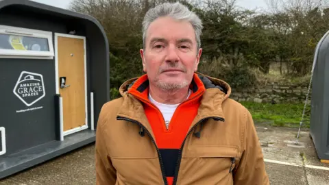 A man with short grey hair, swept back, and a neat short beard and moustache. He is wearing a brown jacket and an orange jumper. He is standing in front of a grey emergency accommodation pod with a wooden door.