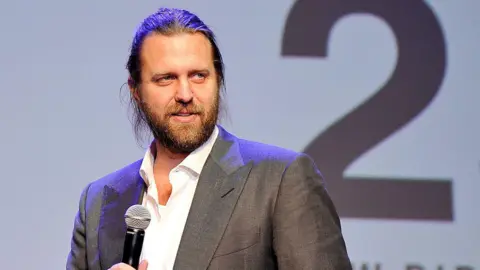 Carl Erik Rinsch seen at an event wearing a sport coat. He holds a mic