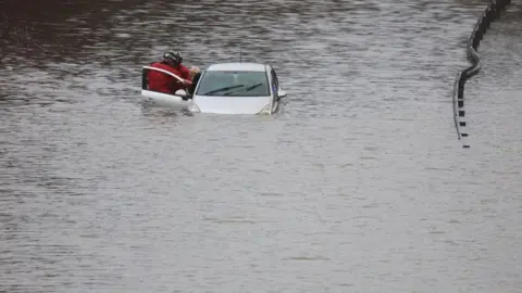 Reuters Firefighters aid a man from his vehicle, after he got trapped in flood waters on the A555 near Bramhall