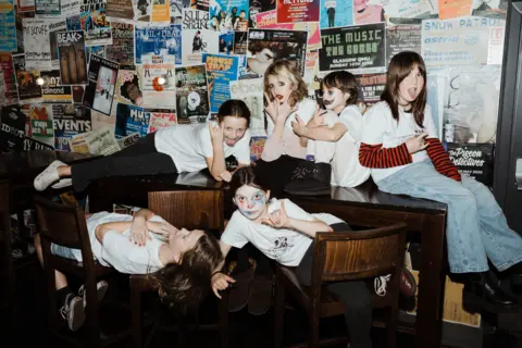 @ellenblairphoto Six girls wear white t-shirts and casual clothes while sitting in various positions on a table an chairs. Behind them you can see lots of layered gig posters. The girls strike poses and two of them have clown make-up on.