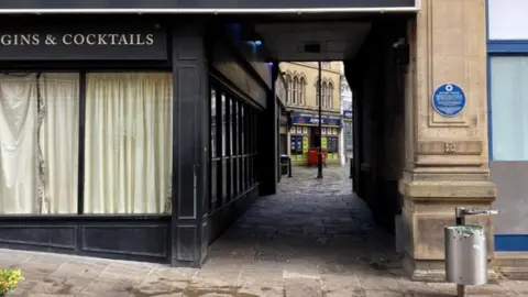 A short alleyway, about 10 metres long. The floor is paved with stone and the area appears clean. A betting shop inside an old stone building can be seen at the other end of the passageway.