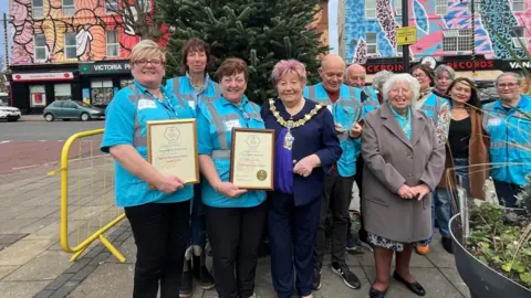 Volunteers wear blue and stand with the mayor holding two awards on a street, next to a planter 