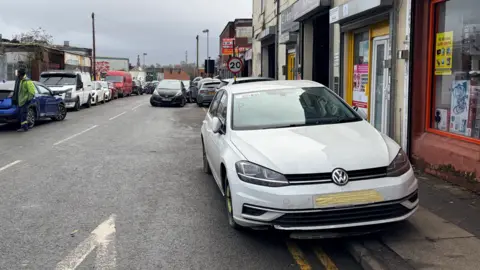 A white car parked on double yellow lines outside a shop.