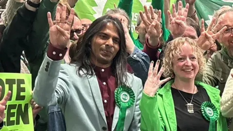 Supporters wearing green rosettes celebrate at a Scottish Green Party event, holding up four fingers as they pose for a group photo in front of green party banners.