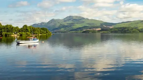 Getty Images A blue and white boat is moored next to a tree-lined shore on Loch Lomond as mountains, a blue sky and clouds are reflected in the water.