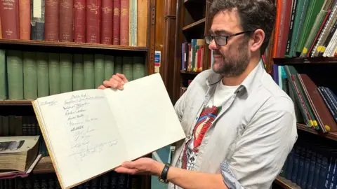 Leeds Central Library A man with brown hair, a brown beard and glasses holds up an open book which has a list of signatures on one side of the page.