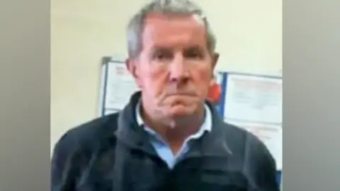 Contributed Richard Scatchard looking at the camera with a stern expression. He is an older man with short grey hair and is wearing a dark coloured fleece over a light blue shirt. The photograph is quite blurry