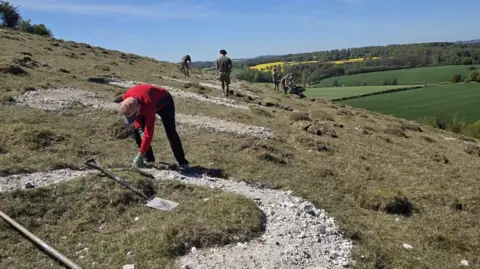 Helen Roberts A man with gloves on leans down and touches a chalk outline of a letter on a grassy hillside.