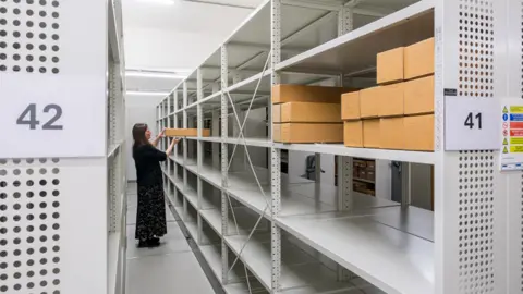 A woman holding a box in a room that has lots of metal shelves in it. There are numbered brown boxes on one of the shelves.