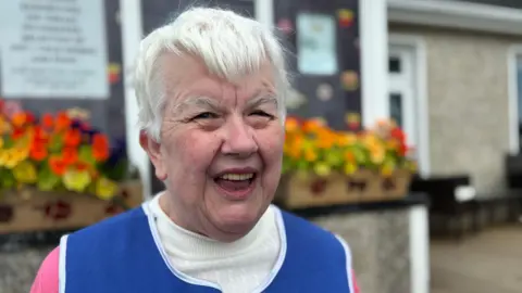 An older woman with short white hair is smiling widely. She is wearing a cream and pink jumper with a blue apron over the top. Behind here there are some window boxes with bright red, yellow and orange flowers.