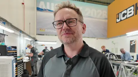 Simon Dowson has short brown hair and glasses and is wearing a dark t-shirt. Behind him is a room of engineers all working on the car.