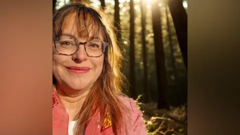 Supplied A woman on the left of the frame is smiling at the camera. She's wearing glasses as has shoulder length brown hair. She is wearing a pink jacket. In the background there is a forest scene.