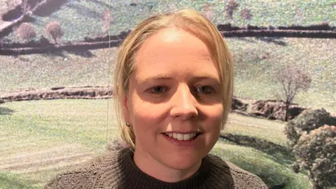 Hannah Crowdy who has blonde hair tied up and is wearing a brown jumper, standing in front of a museum exhibition of fields. 