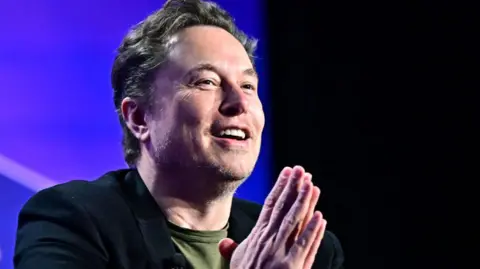 Getty Images Elon Musk laughs during an interview. His palms are clasped together in a prayer position.