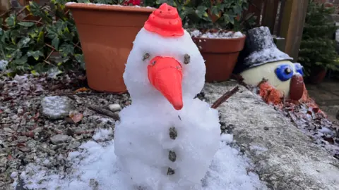 A small snowman with a orange nose and hat. It has two twigs for arms and stones for eyes. There are plant pot behind it.