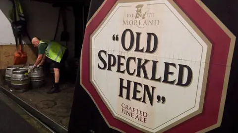 A large Old Speckled Hen beer sign on the side of a lorry, with three beer kegs, and a man bending down, wearing a high-vis jacket. 
