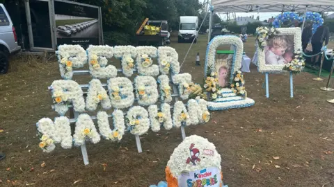 Two photos of a young child surrounded by white, blue and yellow flowers. The floral tributes are in a field. One of the tributes spells out the words "Peter Rabbit Maughan".