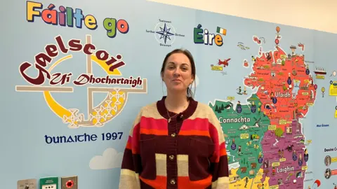 Máire is standing in front of a colourful wall that has a map of Ireland with Irish place names on it. The map is orange, purple, green and yellow. The background of the map is baby blue and then there is writing on the wall in Irish 'Fáilte go', 'Gaelscoil' and 'Eire'. 