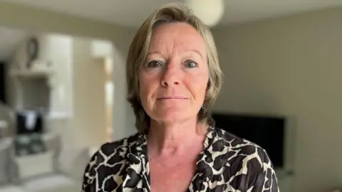 A view of a middle-aged woman looking at the camera for a photo - with a blurred background of a living room.