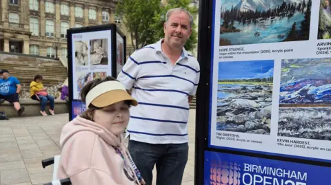 Lily with her dad Graham. They are standing next to her artwork. She has a beige Micky Mouse hoody on and a beige hat. He has a white and blue polo shirt on.