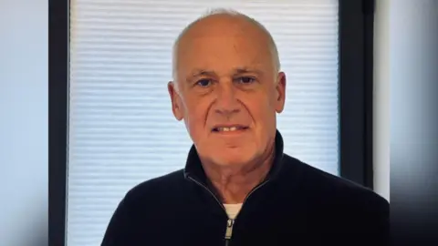 A balding man with grey hair, wearing a black jumper. He is standing inside an office.