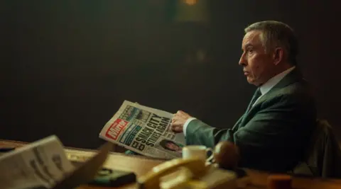 Netflix Steve Coogan, who is wearing a suit and a tie, is sitting on a chair and holding a newspaper as he stares off camera in this still from filming.
