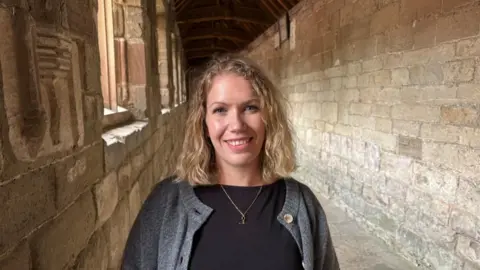 BBC A woman wearing a black top and grey cardigan is smiling at the camera. She has shoulder length blond hair and a gold necklace. She is standing in a stone walled corridor.