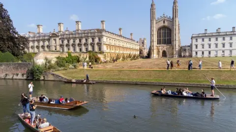 PA Media Four punts on the River Cam along the Backs on a summer's day. Each one has one or two people standing up and steering the flat-bottomed boats. Beyond the river is a shorn meadow running up to King's College Chapel.