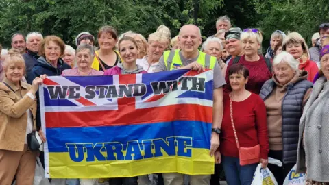 A crowd of people gathered behind four people holding a flag - half the Union Flag and half the Ukrainian flag - which says 'we stand with Ukraine'.