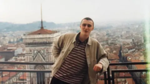 Peter Falconio, a man in his twenties with short dark hair, a beige jacket and hooped shirt. He is standing on a balcony against a backdrop of a city skyline.