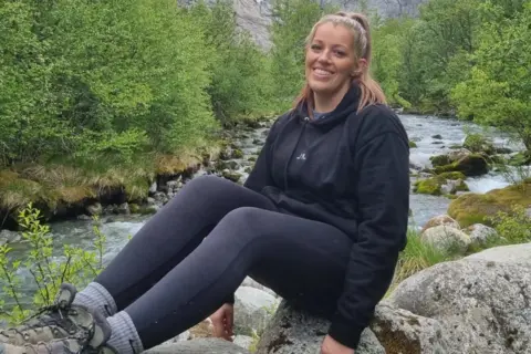Natalie Jones A young woman sits on a rock next to a river with lush greenery in the background. She is smiling into the camera with her hair pulled back, wearing a black hoodie, black leggings and walking boots