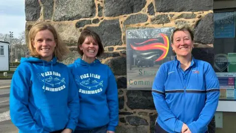 Fizzy Fish Swimmers Three women wearing blue hoodies, with Fizzy Fish written on them, in front of a sign saying Sports Commission.