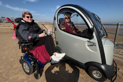 Kevin Shoesmith/BBC Mother and daughter Tracy Lear and Christine Peters, both in mobility scooters, parked in front of the beach at Skegness.