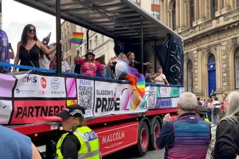 People in colourful clothes and flags on the back of an open-sided lorry that has Pride banners attached 