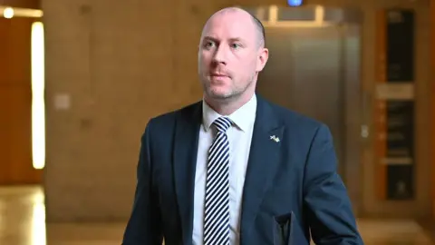 Getty Images Neil Gray, a bald man wearing a dark suit, white shirt and stripy tie, in a medium close-up shot in the Scottish Parliament. 