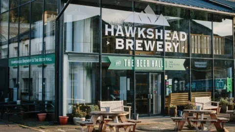 A large building with glass walls with a sign saying "Hawkshead Brewery and the beer hall" on it. There are wooden tables outside.