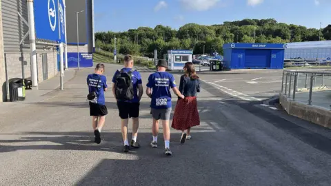 George Carden/BBC Three teenagers in matching blue shirts walking away from the camera outside the Amex stadium in Brighton. They are accompanied by a woman in a denim jacket and red skirt.