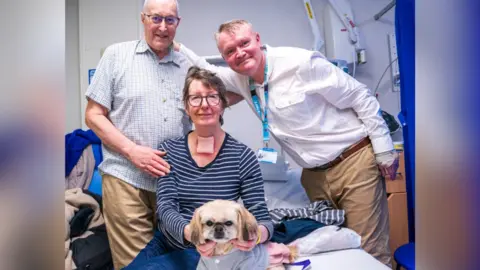 Three people gathered around a hospital bed, with a woman sitting up and holding a small dog dressed in a light outfit, while two men stand beside her. Medical equipment, a blue privacy curtain and folded bedding are visible in the background.