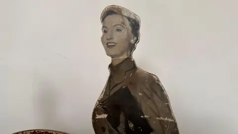 Ben Parker/BBC A close up of the top of the sign owing a woman smiling wearing a jacket and carrying a clutch handbag. 