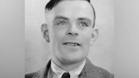 A black and white photo of a smiling Alan Turing - a man with short black hair and wearing a jacket, shirt and tie.