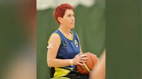 British Wheelchair Basketball Ms Carey playing wheelchair basketball. She wears a blue vest with a yellow design on the arms and sides. It has a number nine in the top left corner and a blue and yellow circle logo in the top right. Ms Carey has the basketball in her hands and is looking towards the direction of the net, away from the camera, with a look of concentration on her face. She has short red hair and plasters on her right upper arm, wrist, and on some fingers. 